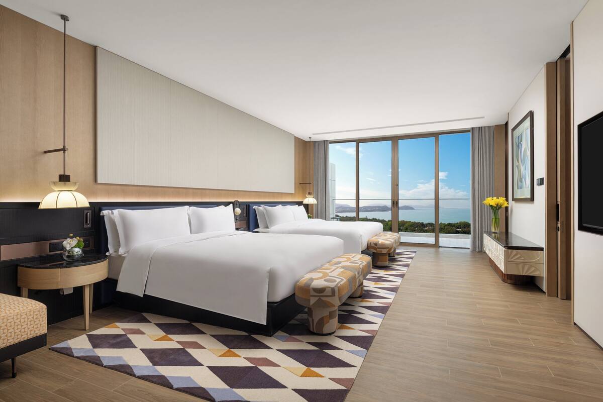 A modern hotel room with two beds, a large window with a city view, a flat-screen TV, and a small side table with a lamp.