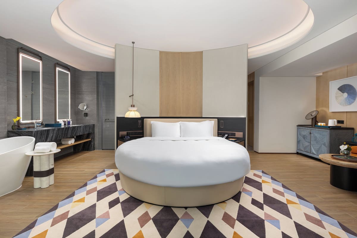 A modern hotel room with a large bed, a round coffee table, a small side table, a large mirror, a bathtub, and a wooden cabinet.