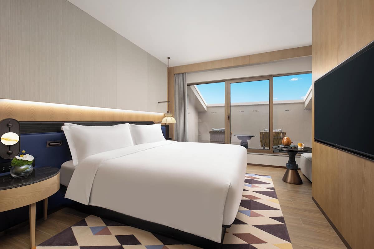 A modern hotel room with a large bed, a desk with a lamp, a small table with a teapot, and a large window with a view of the sky.