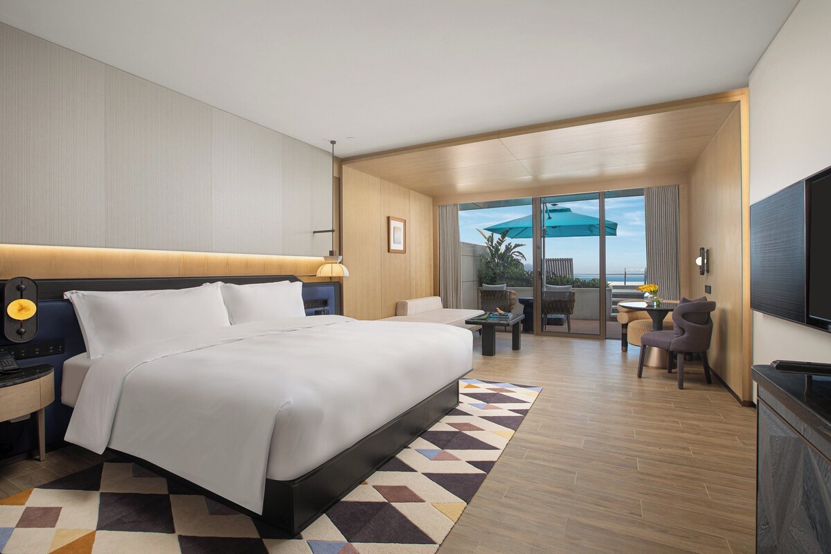 A modern hotel room with a large bed, a desk with a lamp, a TV, and a balcony with outdoor seating.