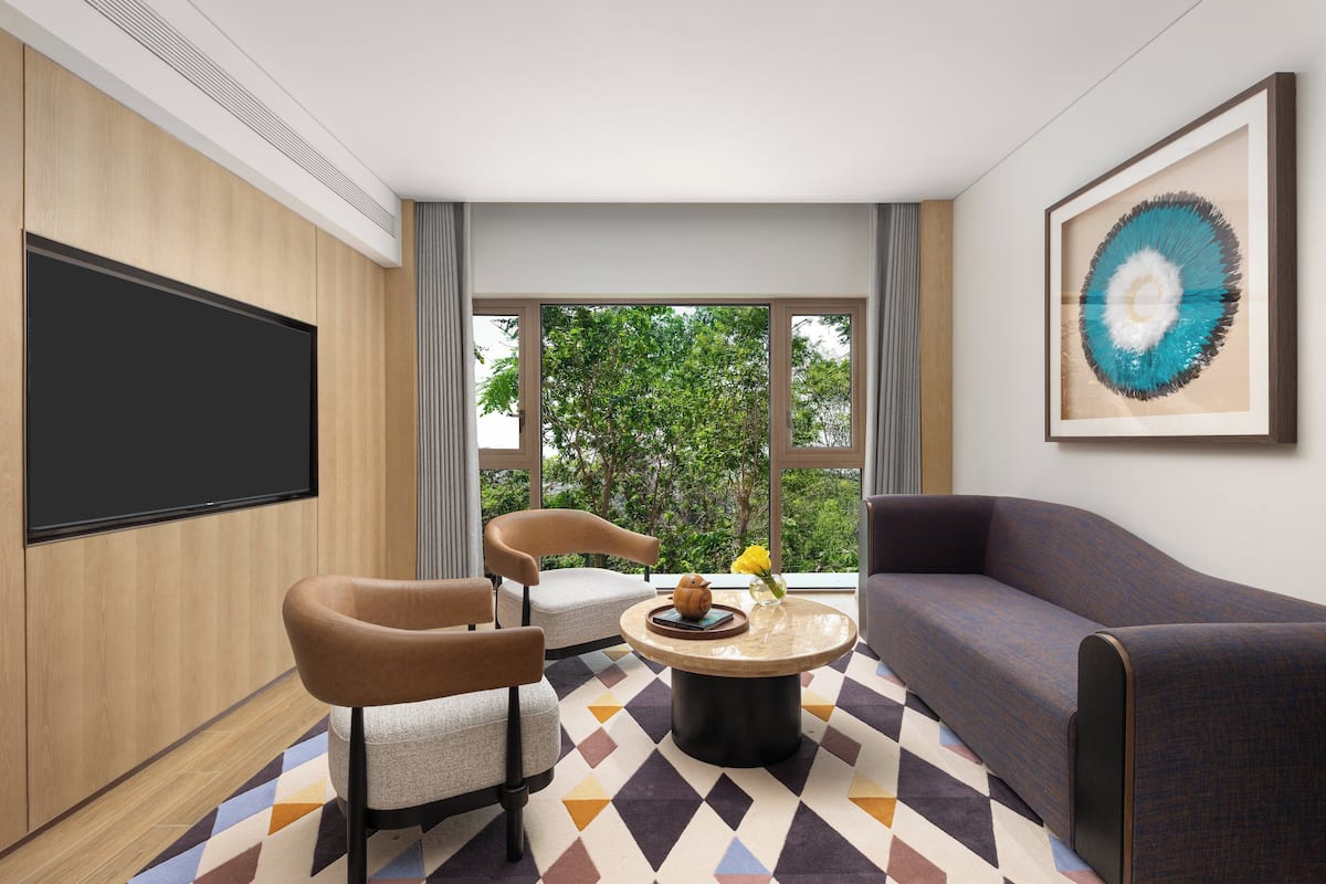 A modern hotel room with a large flat-screen TV, a sofa, two armchairs, a coffee table, and a large window offering a view of trees.