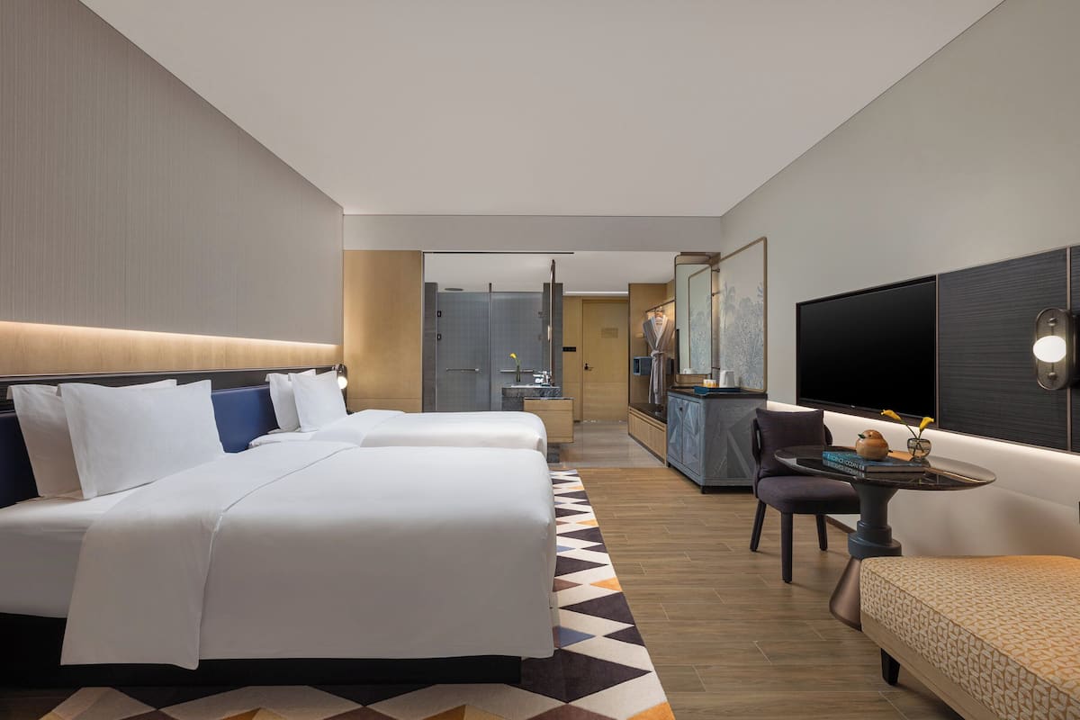 A hotel room with two beds, a desk, a chair, a TV, and a bathroom visible in the background.