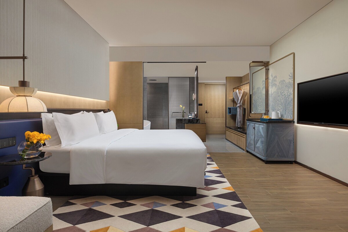 A modern hotel room with a large bed, a flat-screen TV, a bathroom, and a dining area.