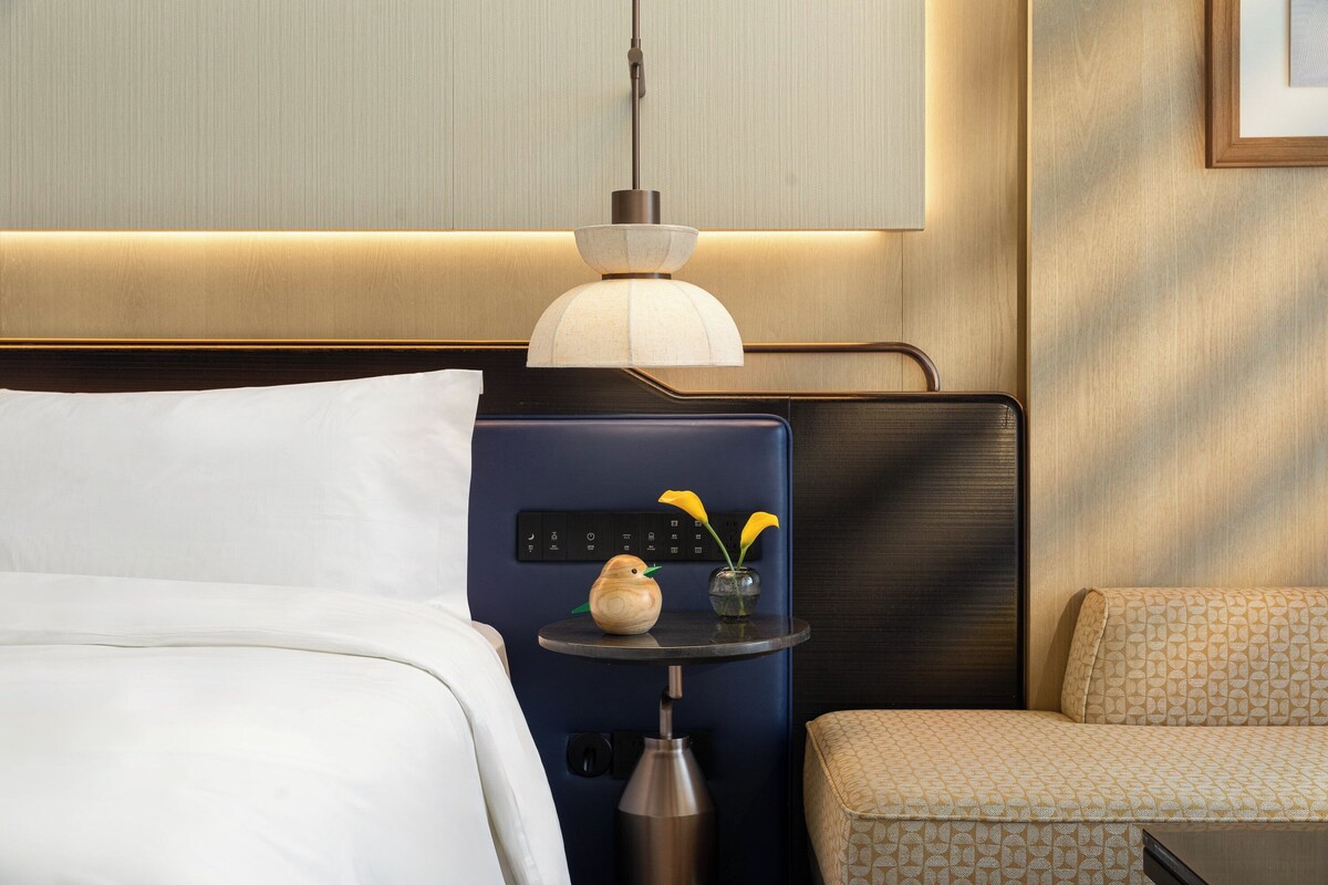 A hotel room with a bed, a bedside table with a lamp, a small vase with flowers, and a sofa.
