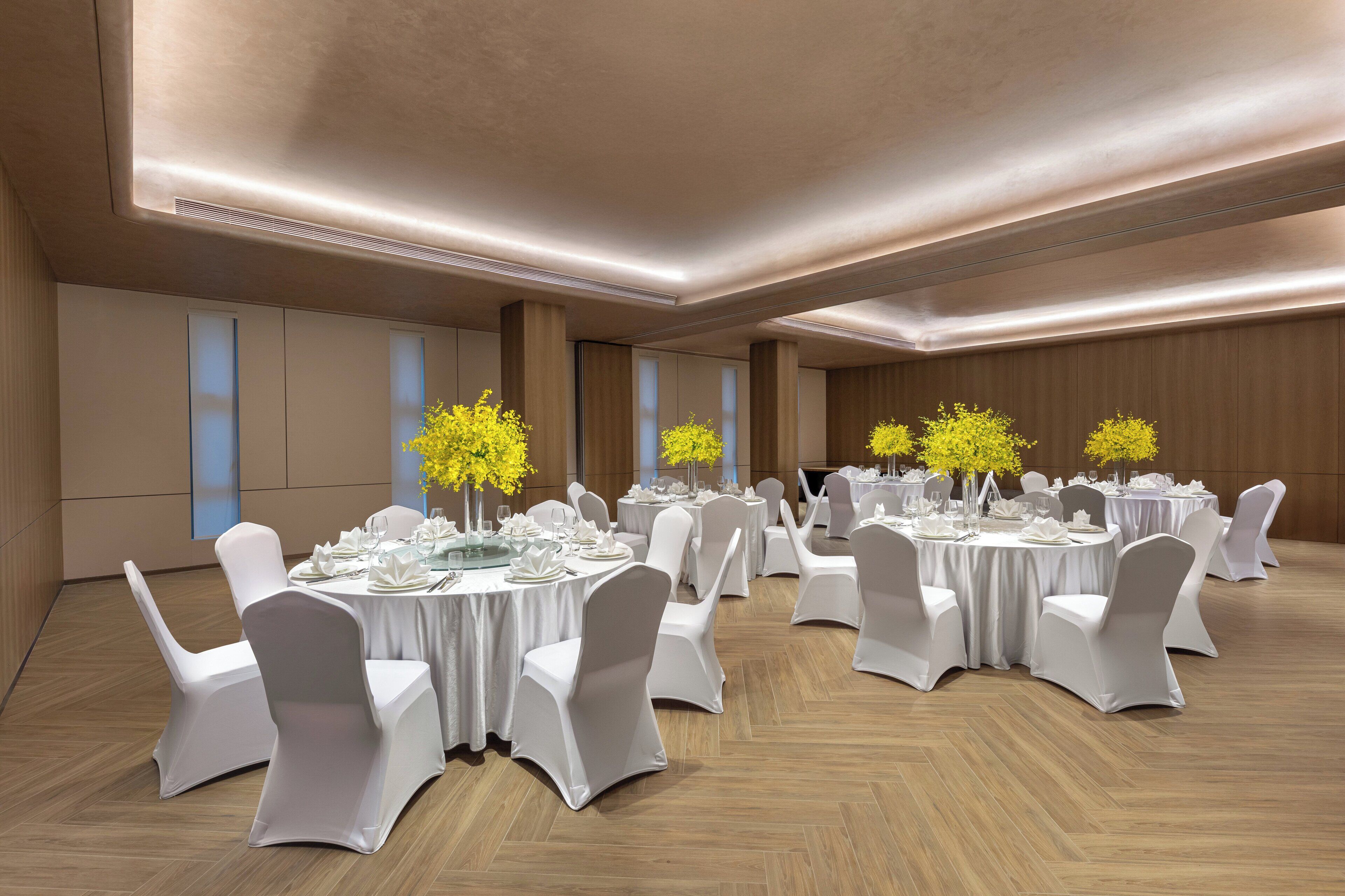 Ein geräumiger Festsaal mit runden Tischen, weißen Stühlen und gelinden floralen Tischdekorationen.