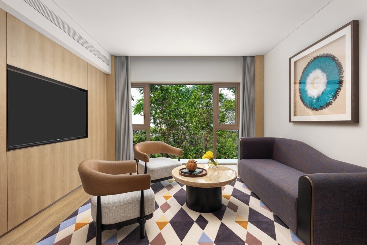 A modern hotel room with a large flat-screen TV, a sofa, two armchairs, a coffee table, and a large window offering a view of trees.