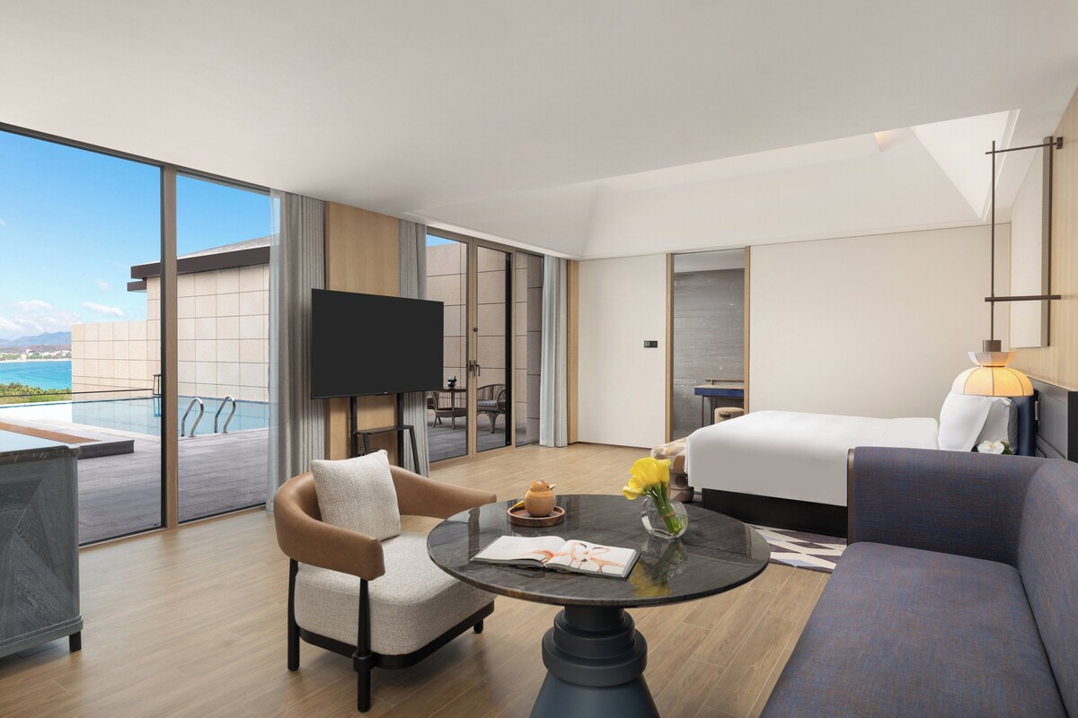 A modern hotel room with a large balcony, a bed, a sofa, a TV, and a dining table with a vase of flowers.