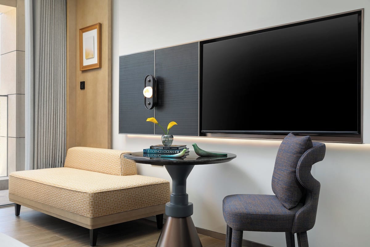 A modern living room with a sofa, a chair, a round table with books and a vase, and a large flat-screen TV mounted on the wall.