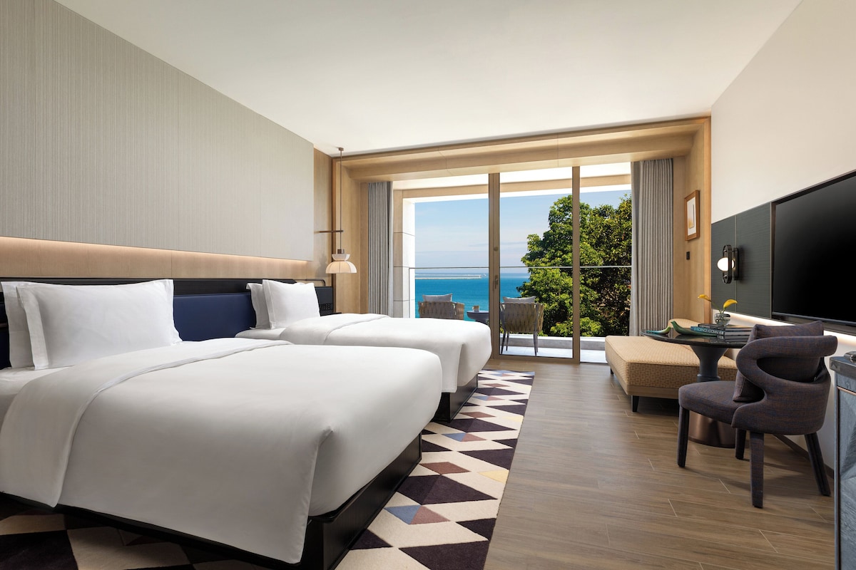 A hotel room with two beds, a television, a desk, and a balcony with a view of the sea.