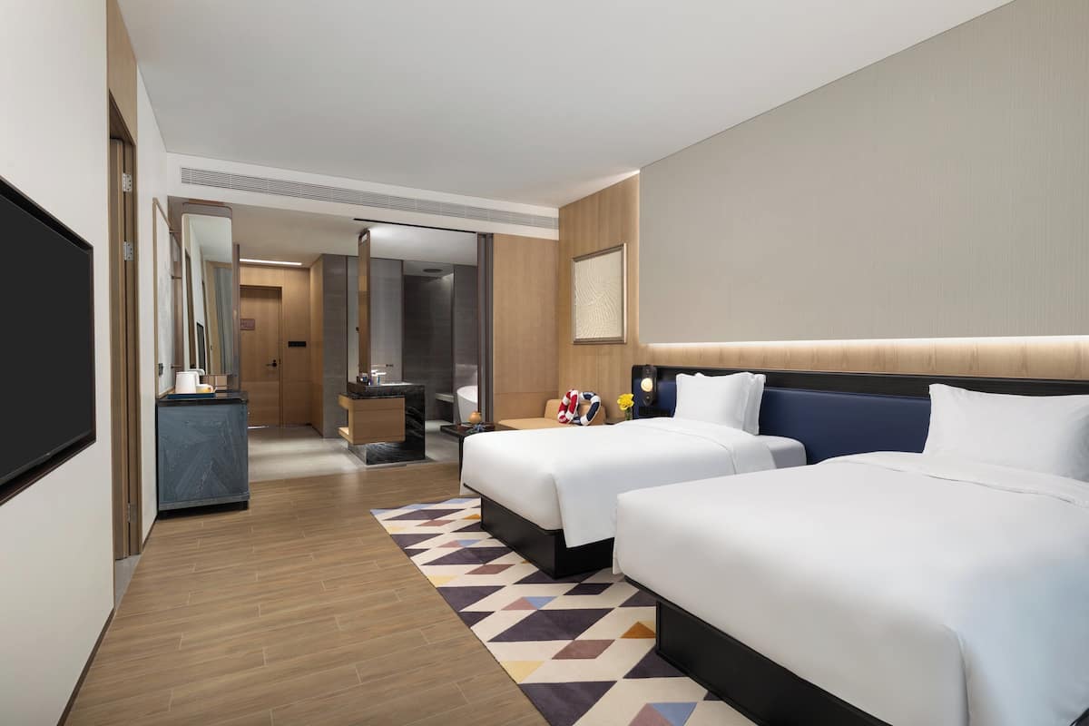 A hotel room with two beds, a desk, a mirror, and a bathroom visible through an open door.