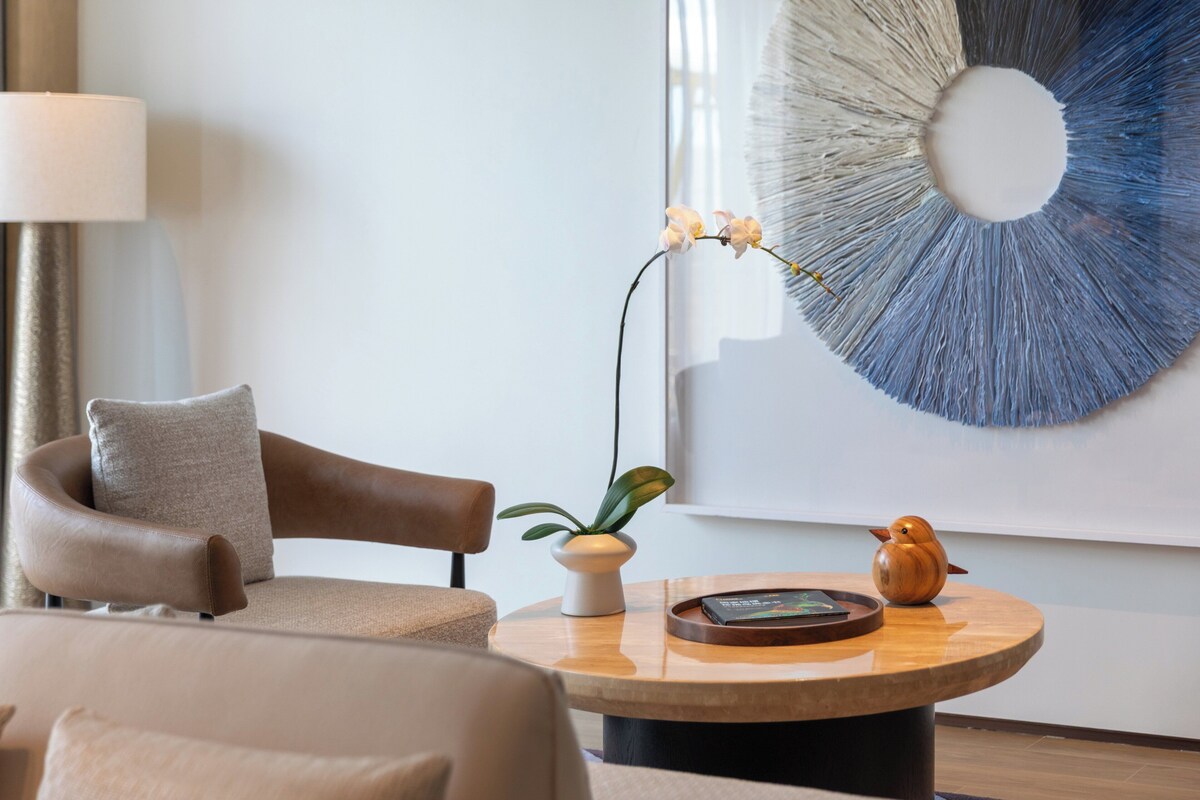 A modern living room with a leather armchair, a round wooden coffee table, a vase with orchids, and a wall art piece.