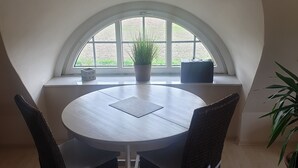 Une table de salle à manger ronde, accompagnée de quatre chaises, d’une plante en pot et d’une fenêtre donnant sur un paysage verdoyant.