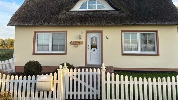 Une maison avec un toit de chaume, une clôture blanche en bois et une boîte aux lettres.