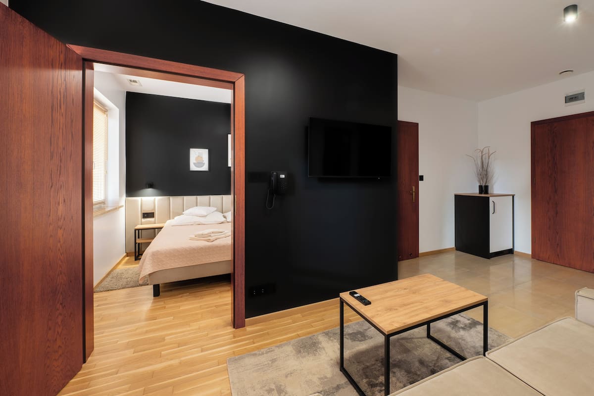 Una habitación de hotel moderna con suelo de madera, una cama con colcha color beige, un televisor negro montado en la pared, una mesita de madera pequeña y un armario.