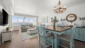 A spacious living area with a large window overlooking the ocean, a dining table with chairs, a television mounted on the wall, and a chandelier. - Edgewater West 72: Beachfront haven for 8 guests! (Gulf Shores)