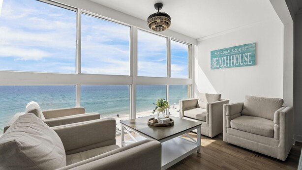 A spacious room with large windows overlooking the ocean, a chandelier, and a sign reading 'Welcome to our Beach House.' - Edgewater West 72: Beachfront haven for 8 guests! (Gulf Shores)