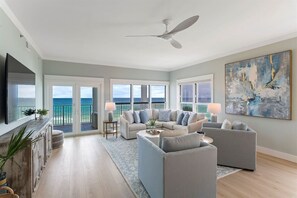 A spacious living room with a large flat-screen TV, a ceiling fan, and a view of the ocean through large windows. - Grand Playa | Gulf Front! | Private Beach! | Pool! (Santa Rosa Beach)