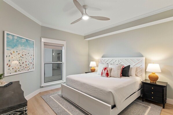 A bedroom with a bed, bedside tables, a ceiling fan, a window, and a painting on the wall.