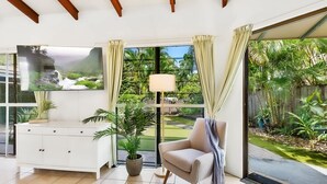 A modern living room with a white cabinet, a beige armchair, and a large window with curtains. - Green Cottage in Trinity Beach (Trinity Beach)