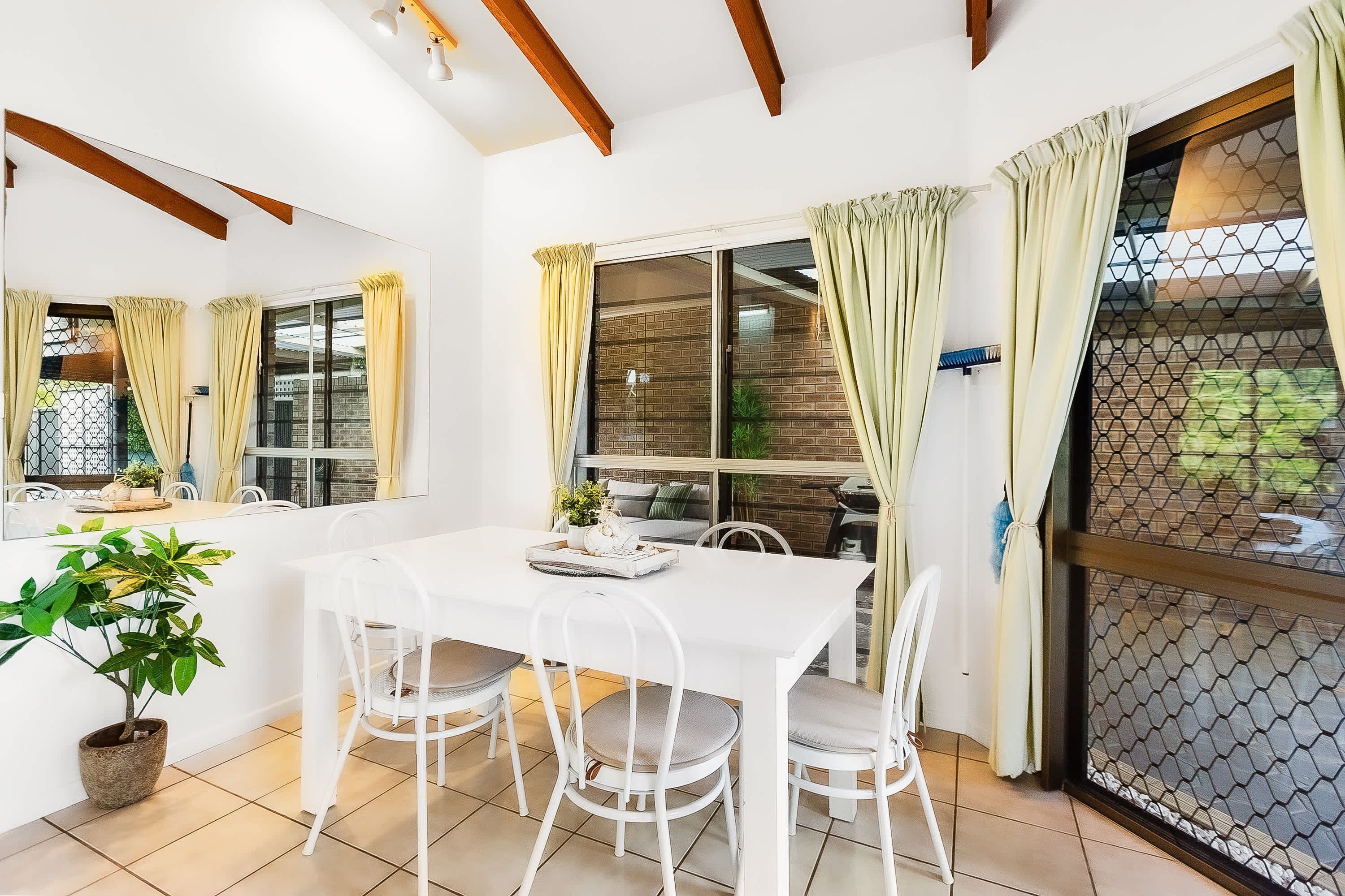 Un comedor con una mesa y sillas blancas, una planta en maceta y vistas a través de una ventana con cortinas.