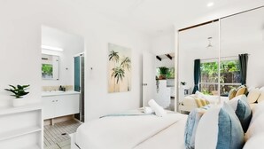 A modern bedroom with a large bed, a bedside table, a wardrobe, a mirror, and a view of a garden through a sliding door. - Green Cottage in Trinity Beach (Trinity Beach)