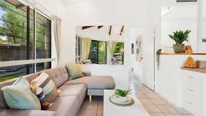 A modern living room with a sectional sofa, a coffee table with a plant, and a kitchen area with white cabinets and a countertop. - Green Cottage in Trinity Beach (Trinity Beach)