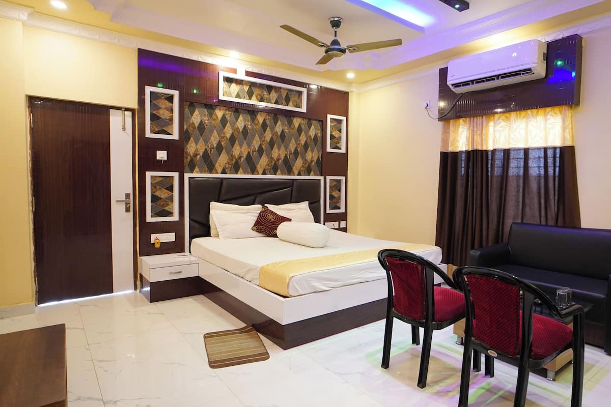 A hotel room with a bed, a desk, two chairs, a ceiling fan, an air conditioning unit, and a window with curtains.