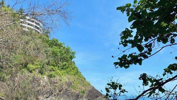Eine Küstenlandschaft mit felsigem Ufer, klarem Wasser und einem Gebäude, das teilweise von Vegetation verdeckt wird.