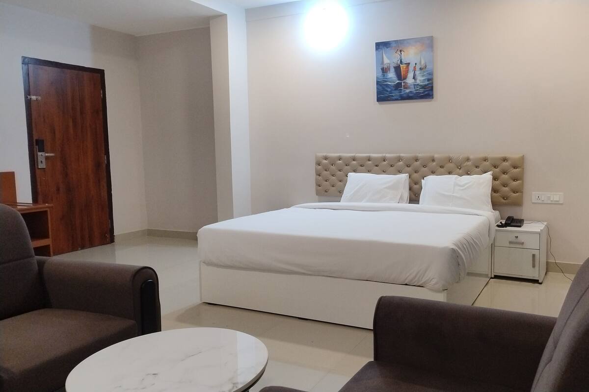 A hotel room with a bed, a nightstand, a chair, a small table, and a painting on the wall.