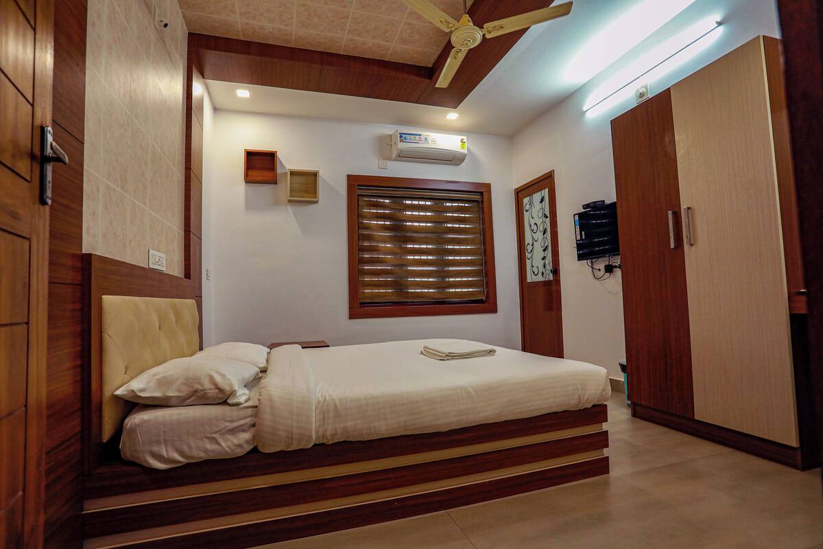 A bedroom with a bed, wooden headboard, a ceiling fan, an air conditioner, a window with blinds, a wardrobe, and a small shelf with a clock.