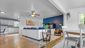 A modern living room with a white sofa, blue cushions, a dining table with chairs, a ceiling fan, and a kitchen in the background.