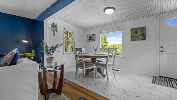 A dining area with a white table and chairs, a potted plant, and a view of the outdoors through large windows.