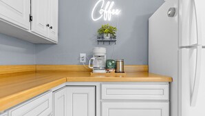 A kitchen with white cabinets, a wooden countertop, a coffee machine, a neon sign, and a shelf with a plant.