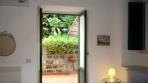 Quarto de hotel com cama, mesa de cabeceira com luminária, um pequeno quadro na parede e vista para um jardim através de uma porta aberta.