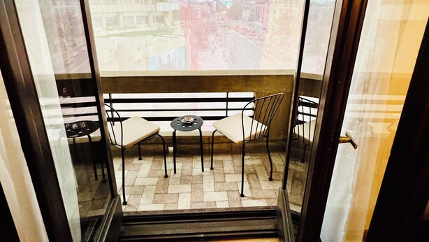 Un balcon doté d’un plancher en parquet à motifs chevrons, une petite table ronde, deux chaises et une vue sur le paysage urbain, visible à travers de grandes fenêtres.