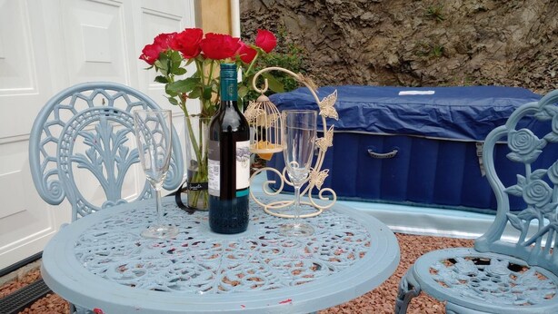 Une table ronde surmontée d’un plateau en métal décoratif, entourée de deux chaises bleues richement ornées. Une bouteille de vin et un verre sont posés sur la table, et un vase de roses rouges se trouve à l’arrière-plan.