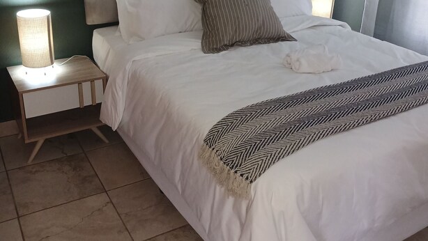 A neatly made bed with a striped pillow, a white pillow, and a white towel, flanked by two bedside lamps on a wooden nightstand. - Rustenburg Cozy Apartment With Private Entrance & BBQ (Rustenburg)