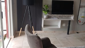A modern living room with a grey armchair, a flat-screen TV, a floor lamp, and a glass coffee table. - Rustenburg Cozy Apartment With Private Entrance & BBQ (Rustenburg)