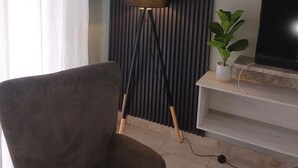 A living room with a dark-colored sofa, a floor lamp, a television on a stand, and a potted plant. - Rustenburg Cozy Apartment With Private Entrance & BBQ (Rustenburg)