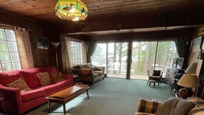A cozy living room with a red sofa, a plaid sofa, and a wooden coffee table. There is a large window with curtains, a ceiling light fixture, and a view of the outdoors.