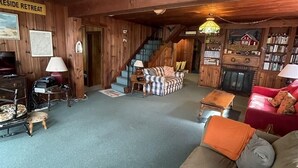A cozy cabin interior with a wooden floor, a sofa, a red chair, a staircase, and a bookshelf.
