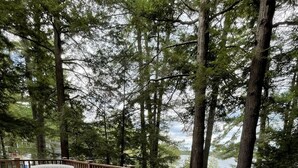 A wooden deck with white plastic chairs and a table, surrounded by tall trees and overlooking a body of water.