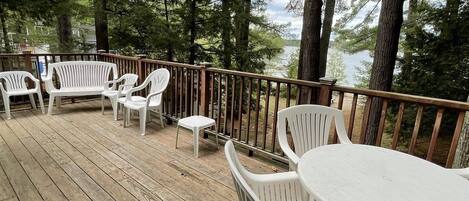 A wooden deck with white plastic chairs and a table, surrounded by tall trees and overlooking a body of water.