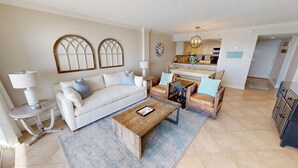 A living room with a sofa, coffee table, and armchairs, a dining area with a table and chairs, and a kitchen in the background. - Beach House 301a 2 Bedroom Condo (Miramar Beach)