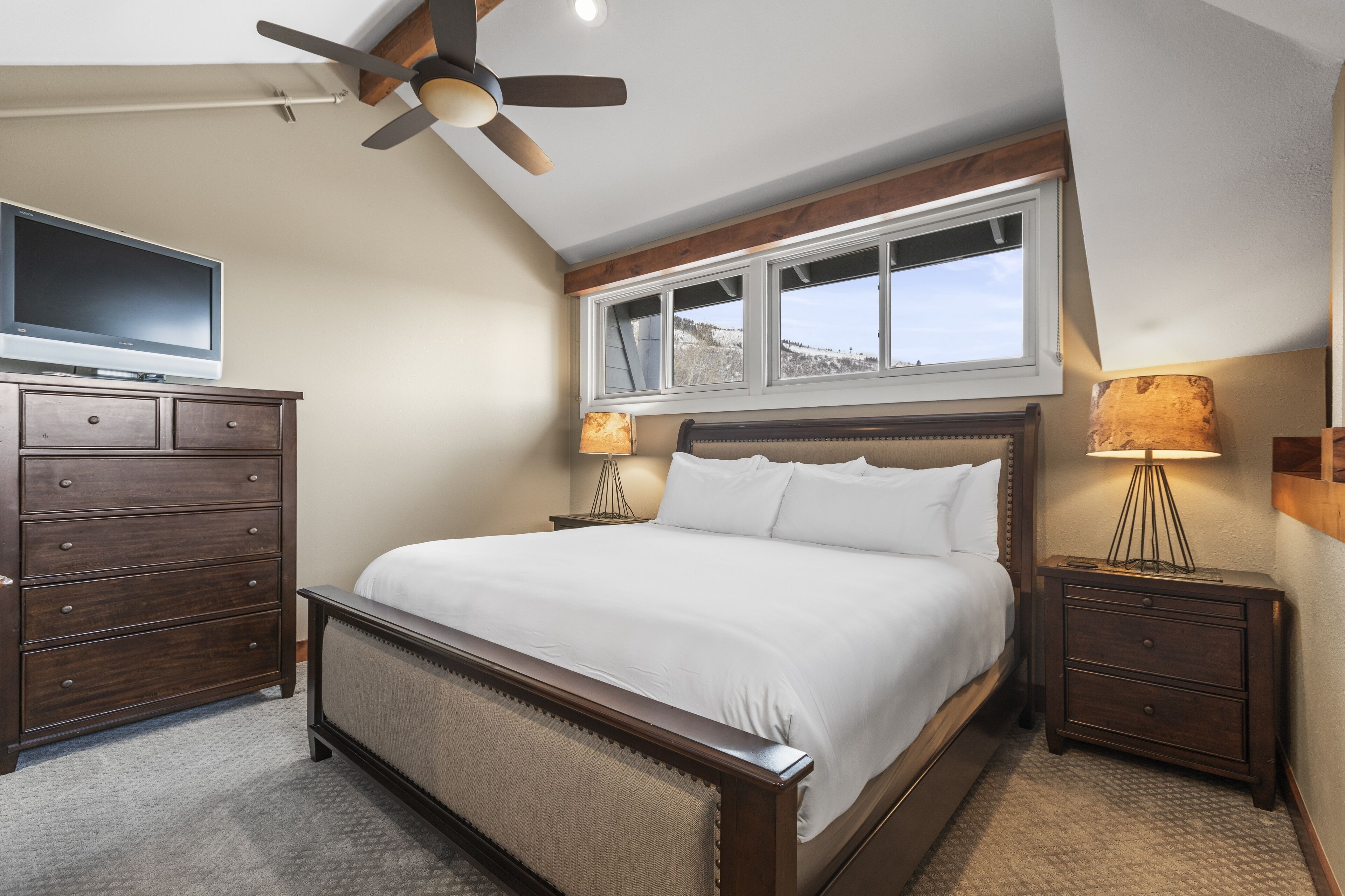 A bedroom with a large bed, wooden nightstands, a ceiling fan, a TV, and a window with a view of a snowy landscape.