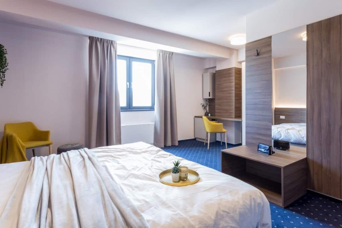 A hotel room with a bed, a desk, a chair, a small table with a tray, and a window with curtains.