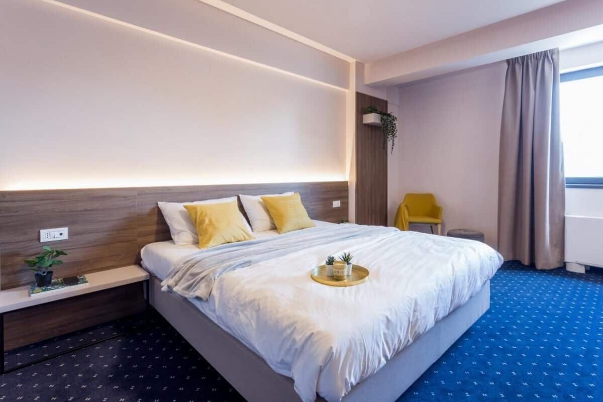 A hotel room with a large bed, a bedside table with a plant, a tray with small plants, and a window with curtains.