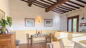 Ett mysigt vardagsrum med ett träbord, en soffa och en matplats med stolar. På väggen hänger inramade tavlor och det finns ett fönster med gardiner.