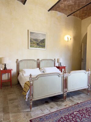 A bedroom with a large bed, a nightstand, a lamp, a painting on the wall, and a patterned rug. - The Little House (Toscana)