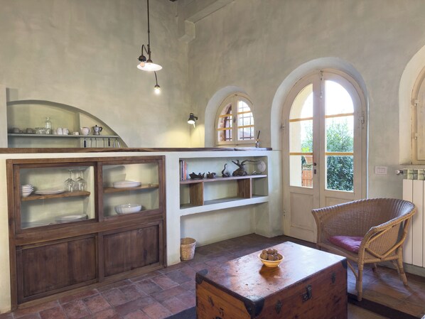 A rustic kitchen with wooden cabinets, a wicker chair, and a view through an arched window.
